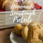 Two finished pretzels on a white plate in front of rolls on a red scarf in a ceramic basket Pinterest banner.