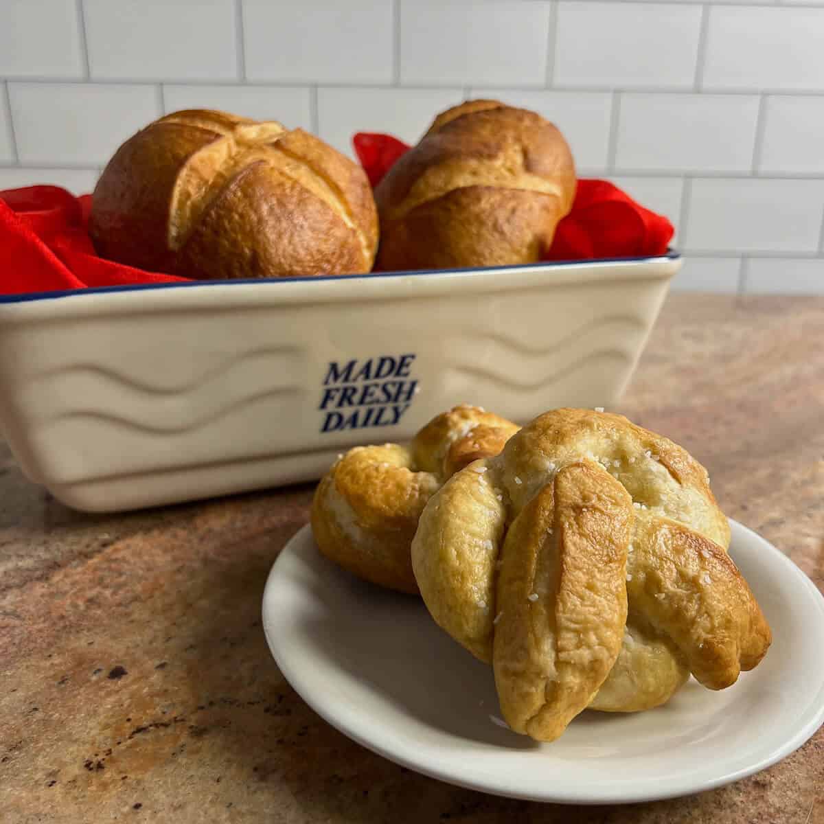 Two finished pretzels on a white plate in front of rolls on a red scarf in a ceramic basket.