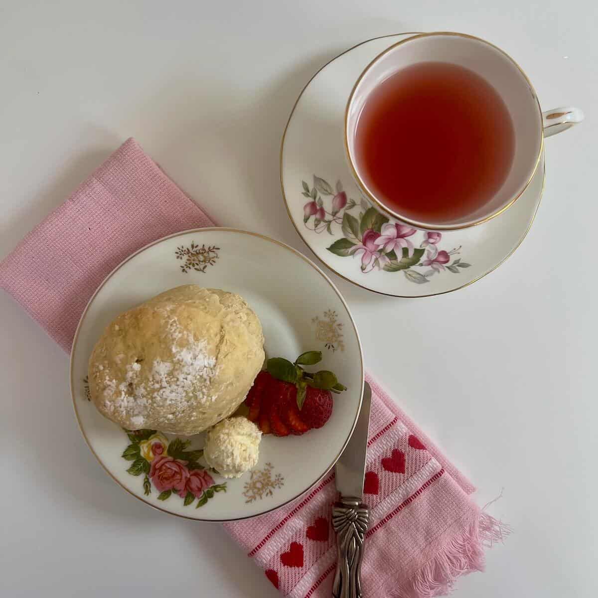 Clotted cream alongside a scone & cut strawberry on a flowered plate atop a pink towel with a small spreader next to a teacup from overhead.