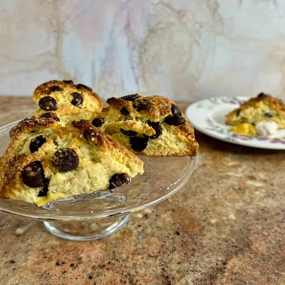 Chocolate chip scones on a cake stand with one on a flowered plate behind.