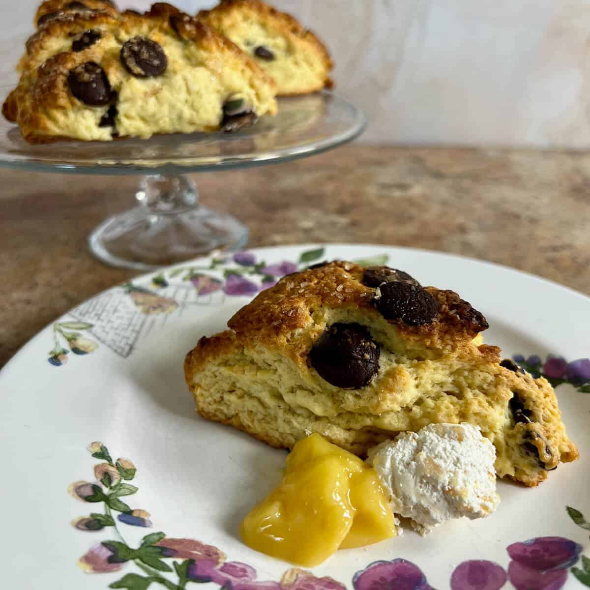 Chocolate chip scones on a flowered plate beside lemon curd & clotted cream with more on a cake stand behind.