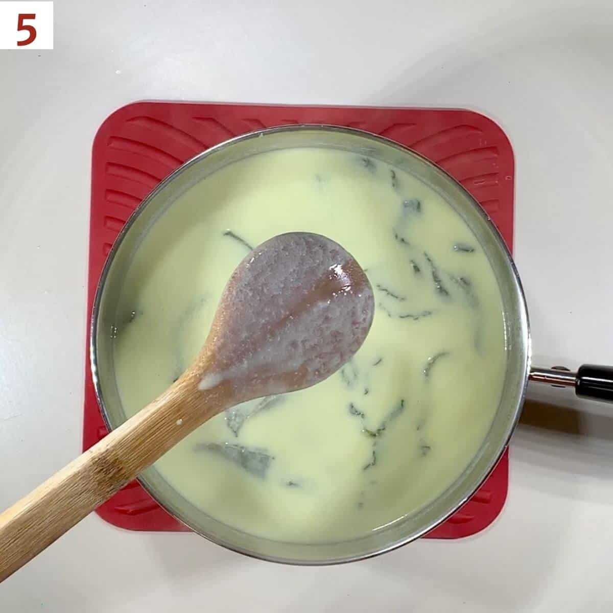 Wooden spoon checking thickness of cooked custard in pot below.