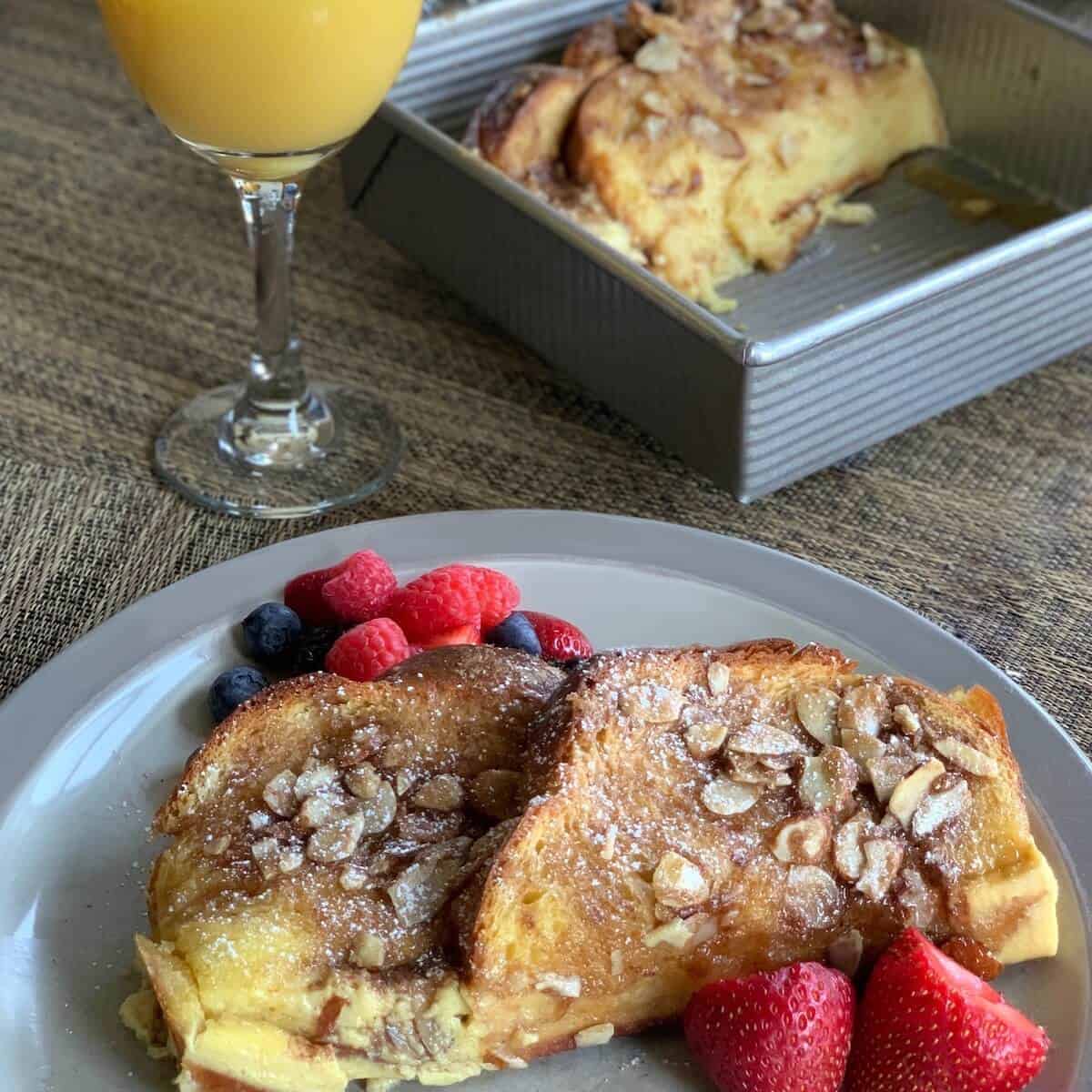 French Toast with berries on a grey plate with orange juice & pan behind.