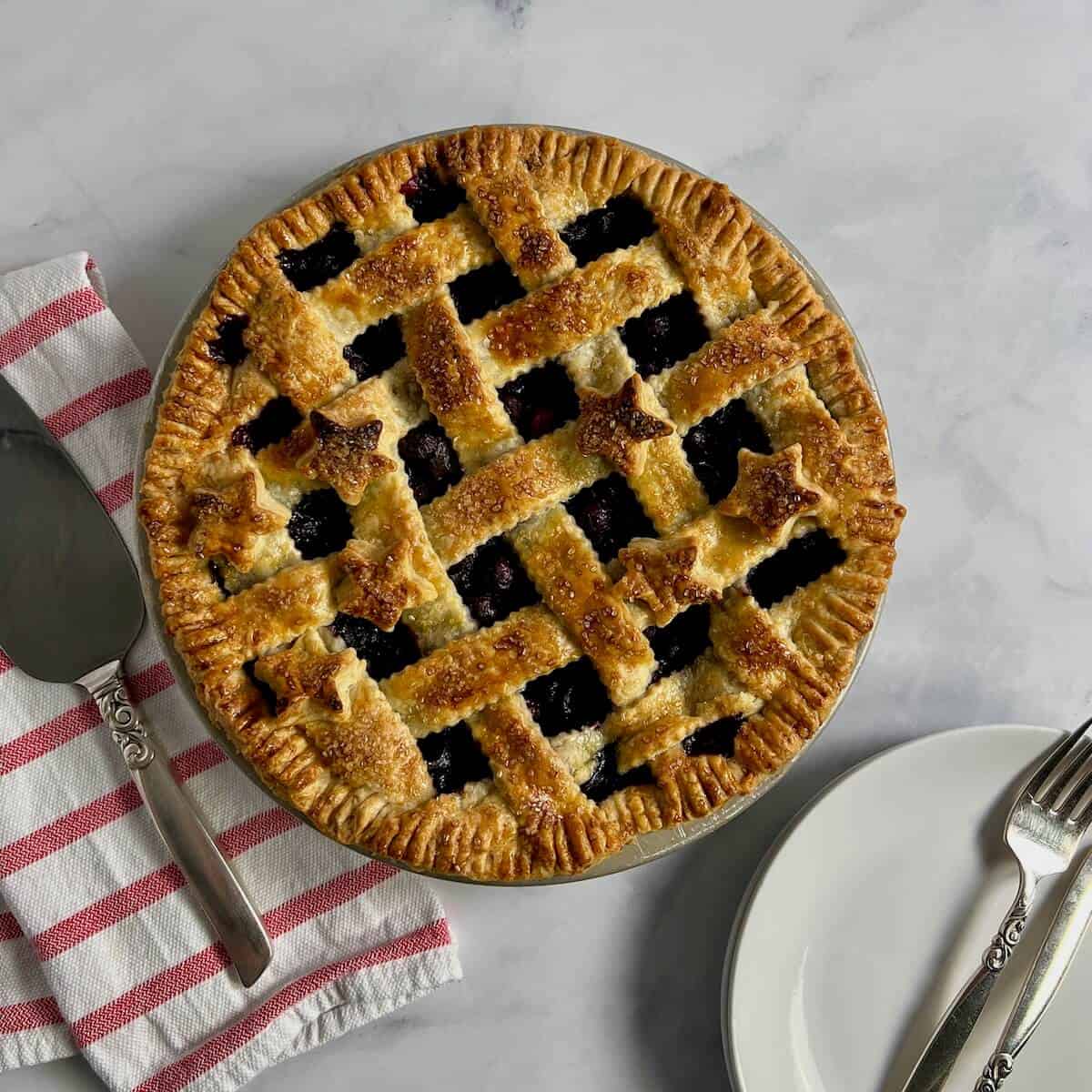 Lattice blueberry pie on a red & white striped towel next to a pie server & white plates with forks from overhead.