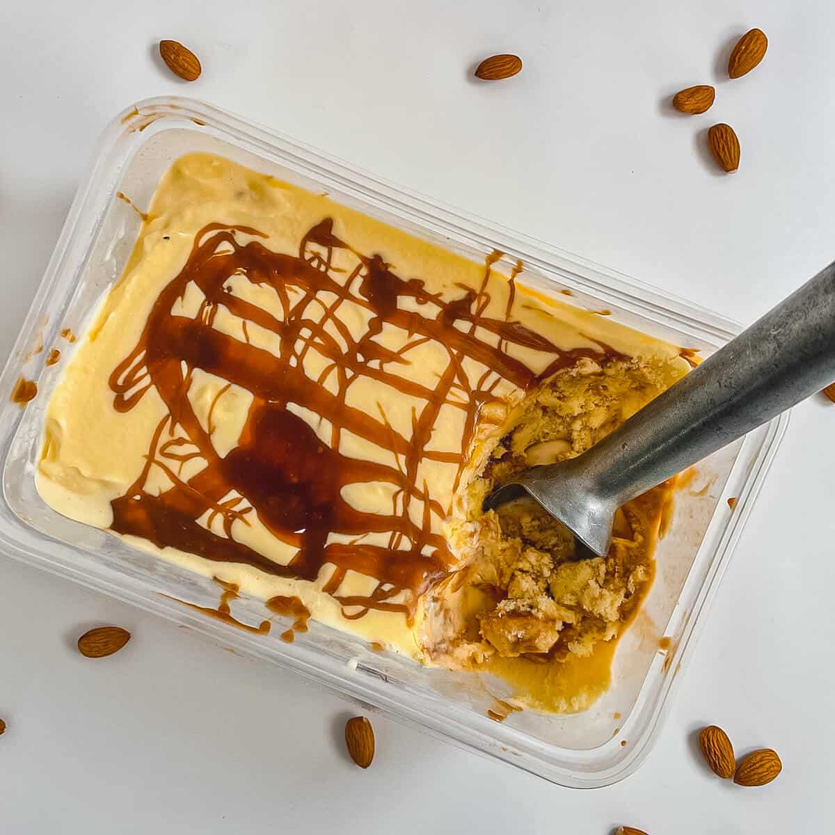 Tub of Caramel Swirl Almond Gelato drizzled with caramel sauce with a scooper & almonds from overhead.
