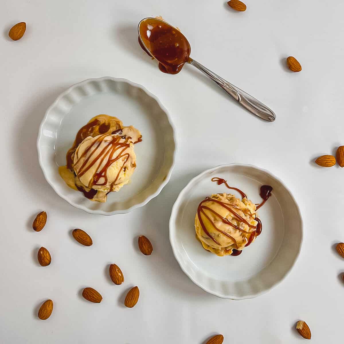 One scoop of Caramel Swirl Almond Gelato drizzled with caramel sauce in two white bowls next to a spoon & almonds from overhead.