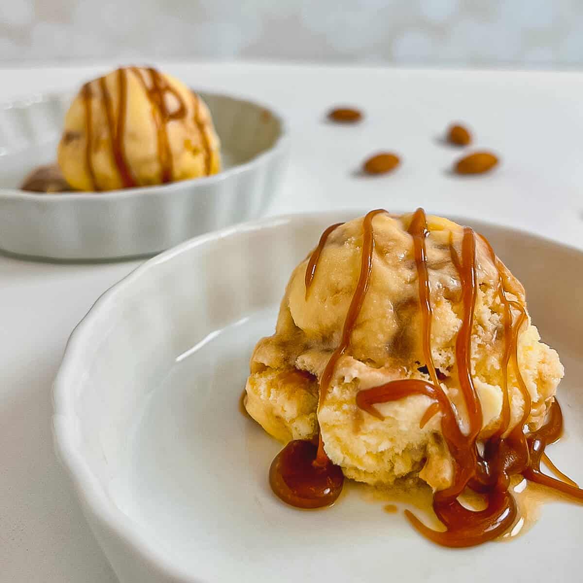 One scoop of Caramel Swirl Almond Gelato drizzled with caramel sauce in a white bowl with another bowl behind.