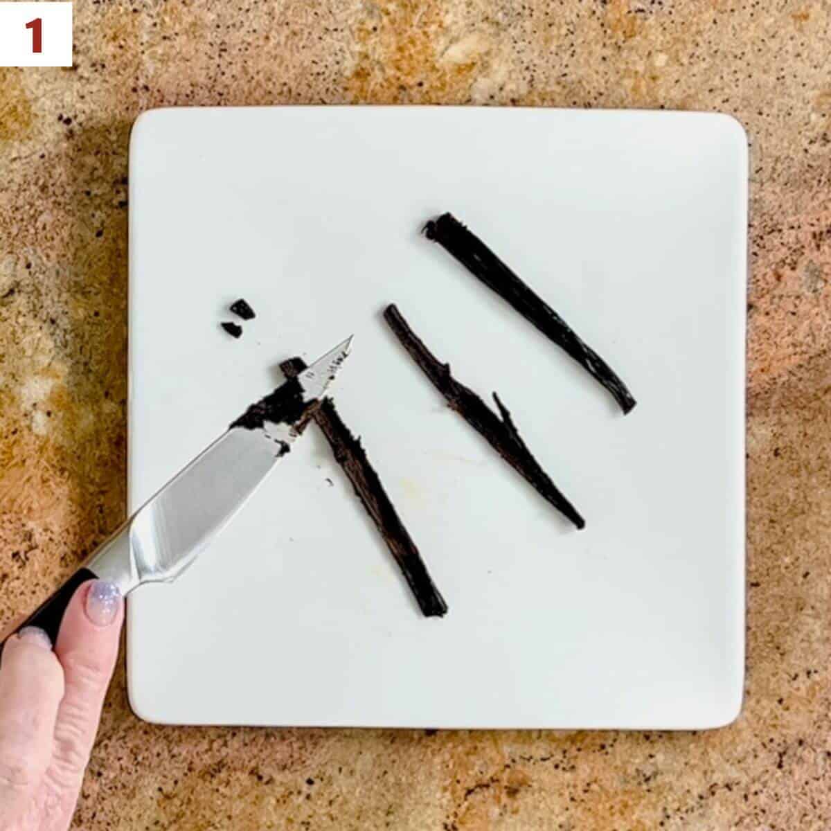 Scraping the seeds from a split vanilla bean pod on a white square plate.