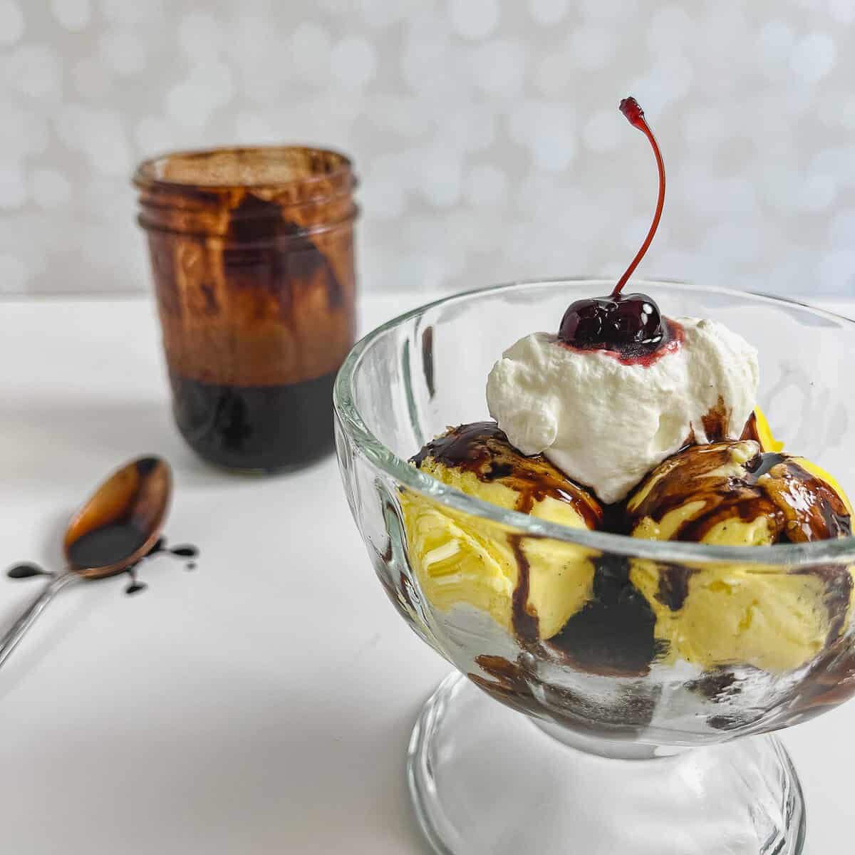 French vanilla ice cream sundae in front of jar of chocolate syrup and a spoon.