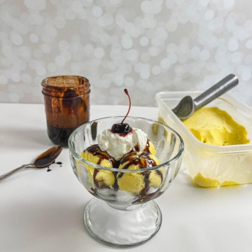 French vanilla ice cream sundae in front of jar of chocolate syrup, spoon, and tub of ice cream with an ice cream scoop.