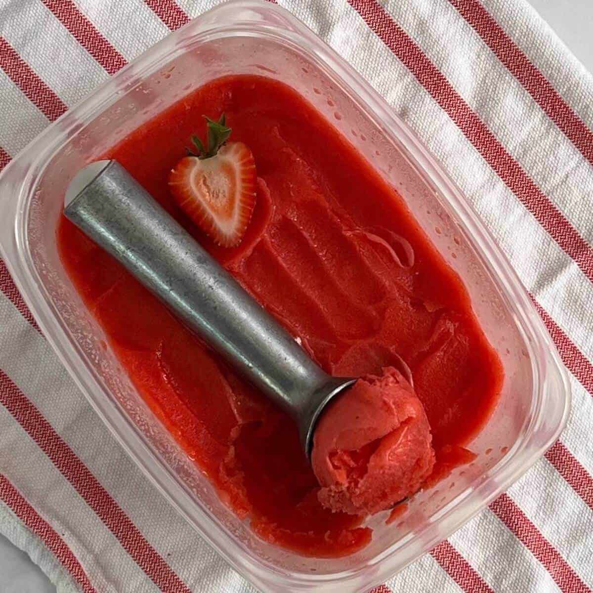 Tub of strawberry mint sorbet with a ice cream scoop on a red & white striped towel from overhead.