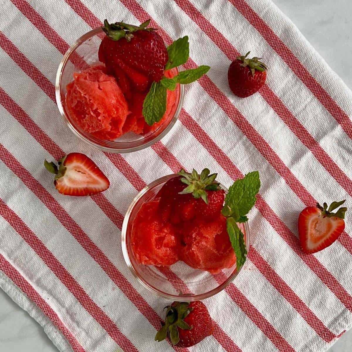 Two bowls of strawberry mint sorbet garnished with strawberries & mint on a red & white striped towel from overhead.