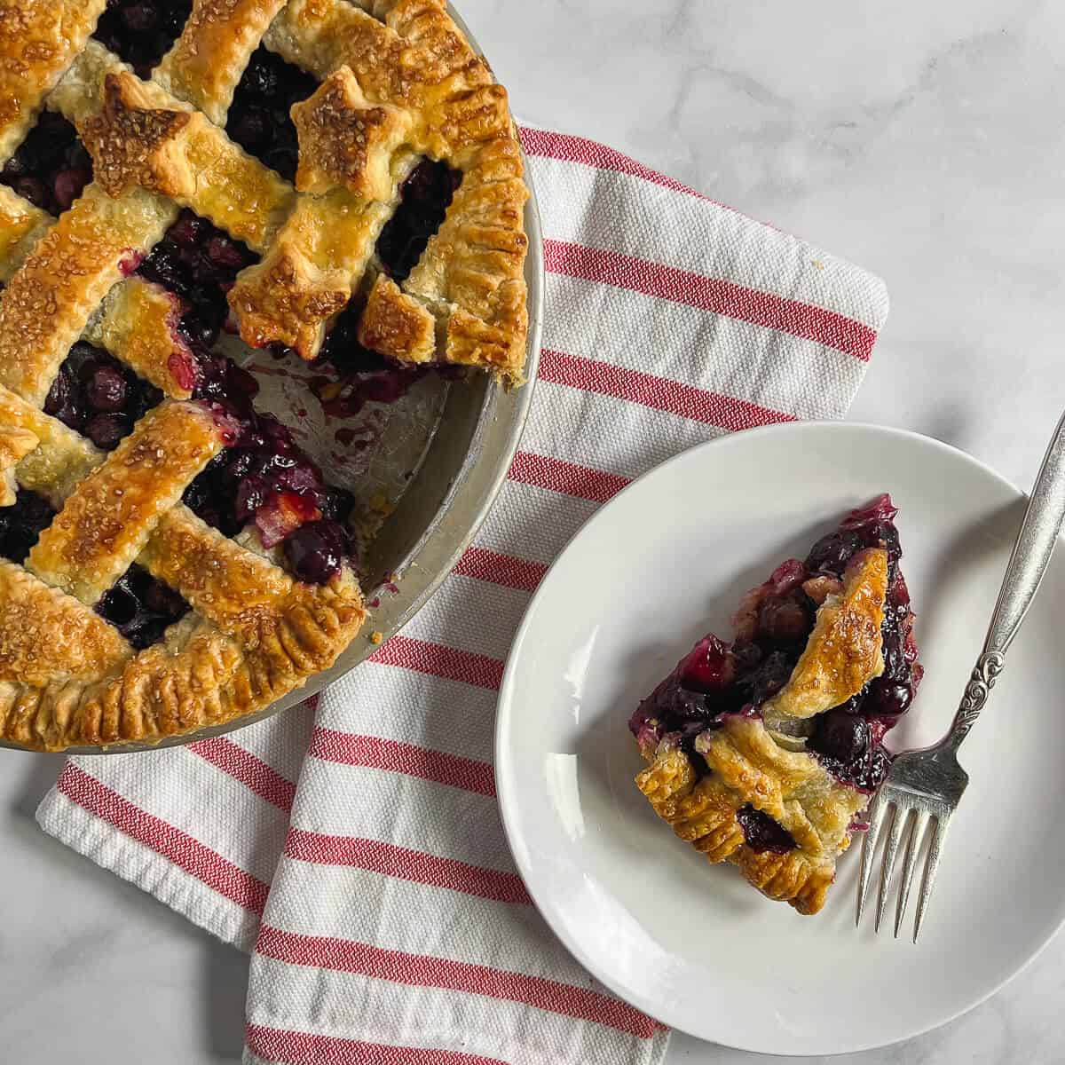 Slice of blueberry pie on a white plate with a fork next to a baked pie with a slice missing on a red & white striped towel from overhead.