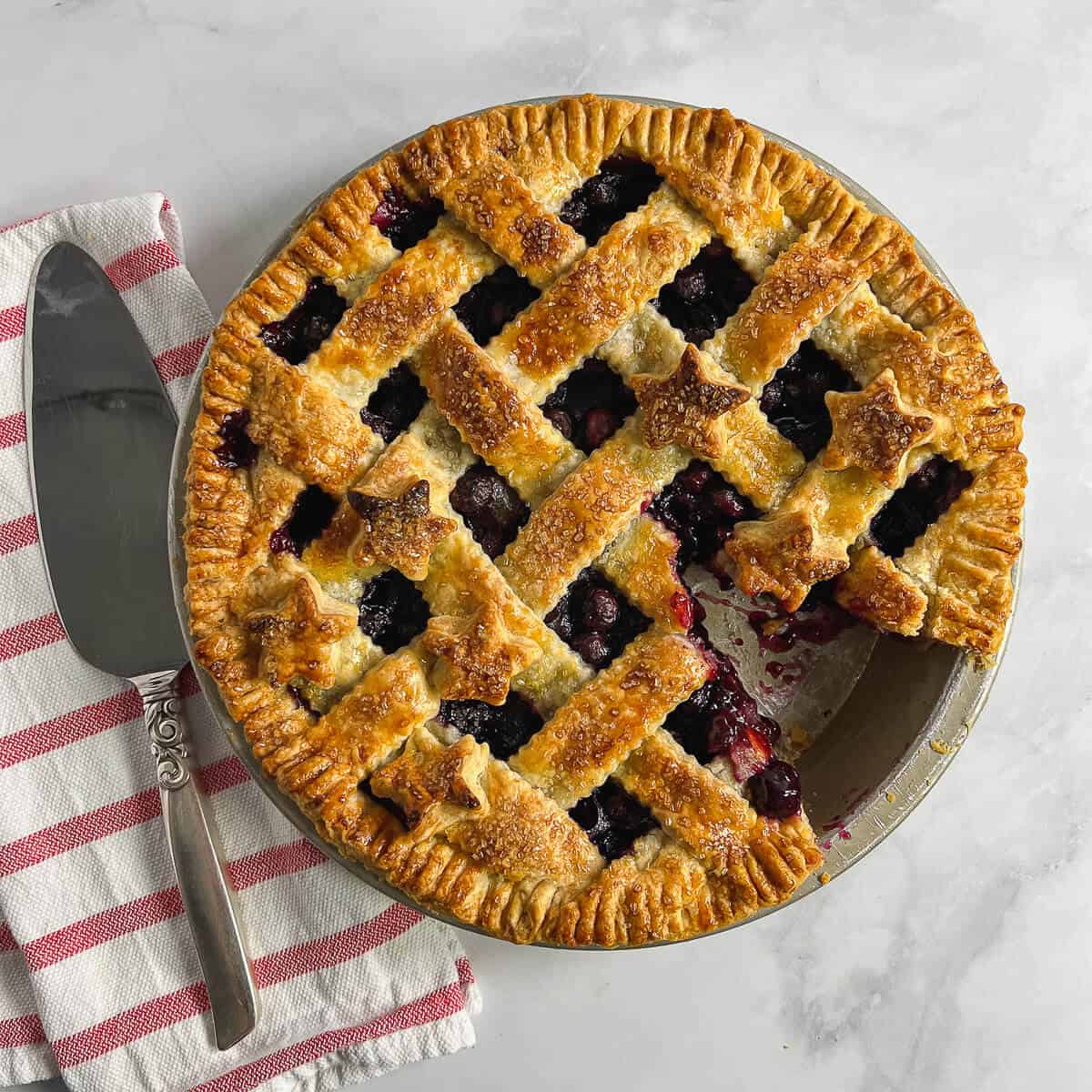 Baked blueberry pie with slice missing next to a pie slicer on a red & white striped towel from overhead.