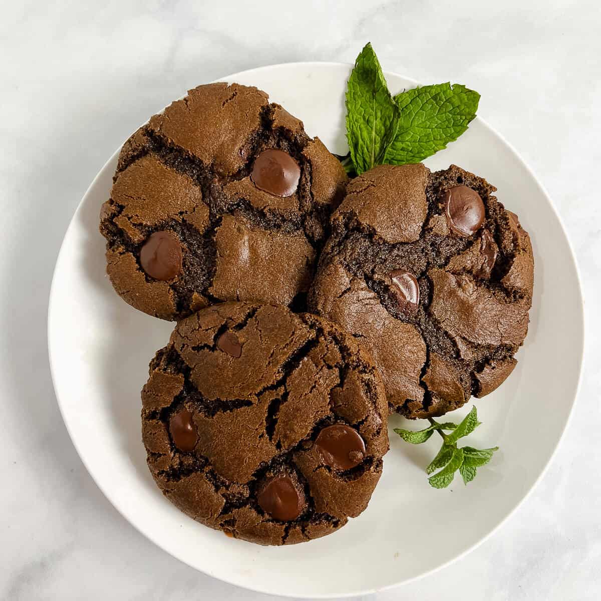 Three Double Chocolate Mint Cookies on a white plate with mint from overhead.