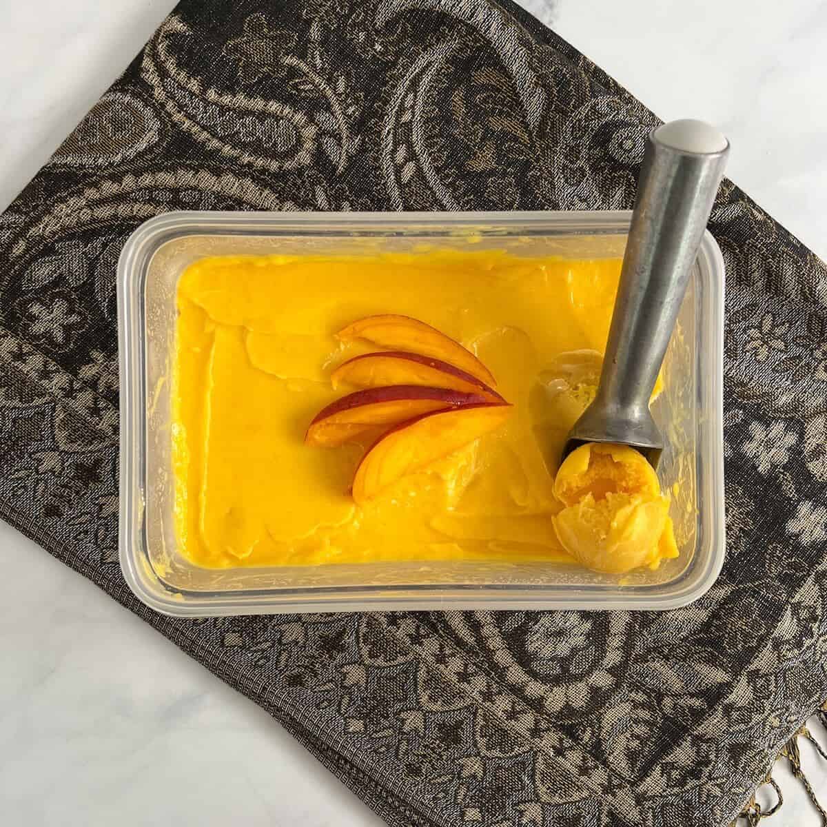 Tub of Mango Sorbet garnished with mango slices and an ice cream scoop on a brown patterned scarf from overhead.