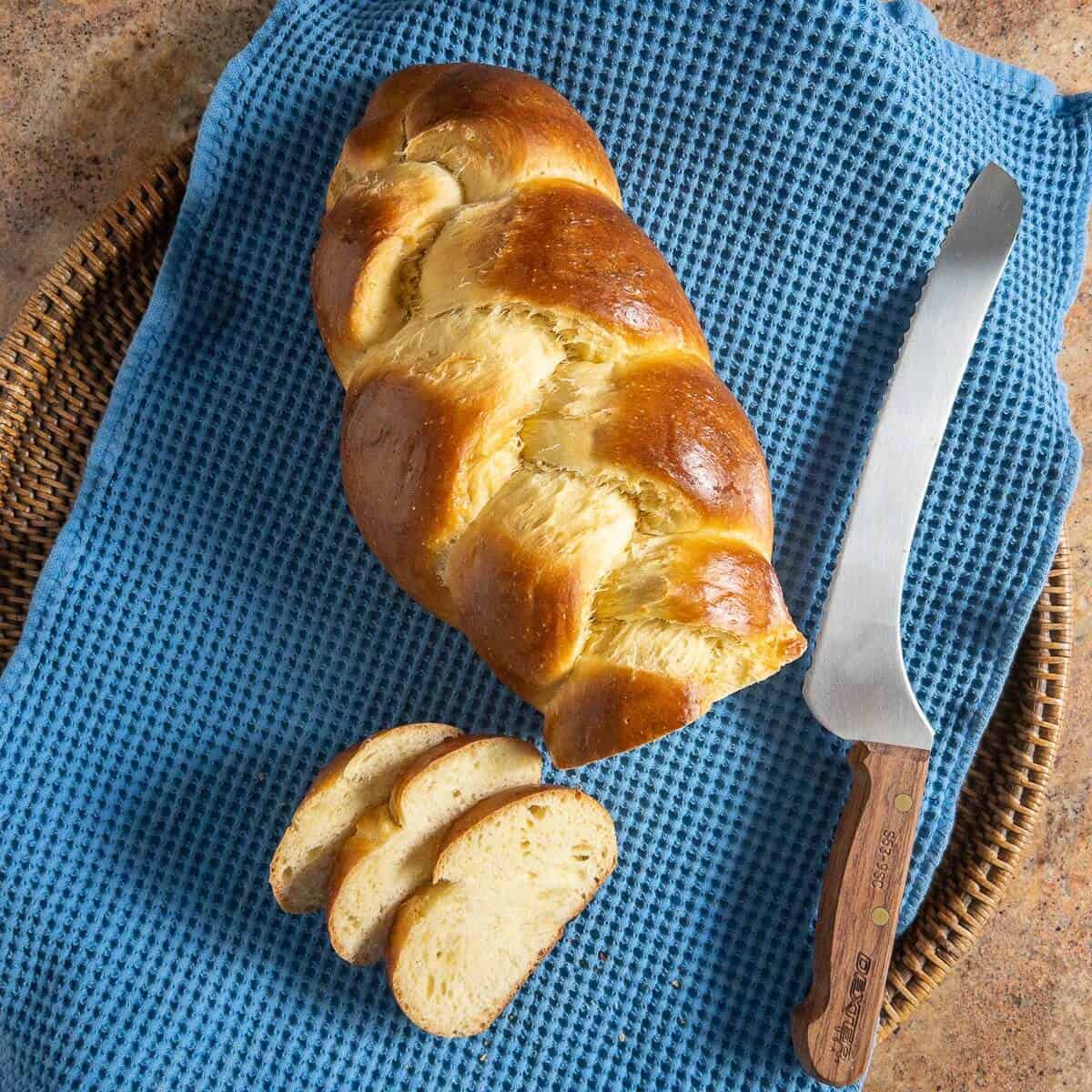Sliced challah loaf on a blue towel next to a bread knife viewed from overhead.