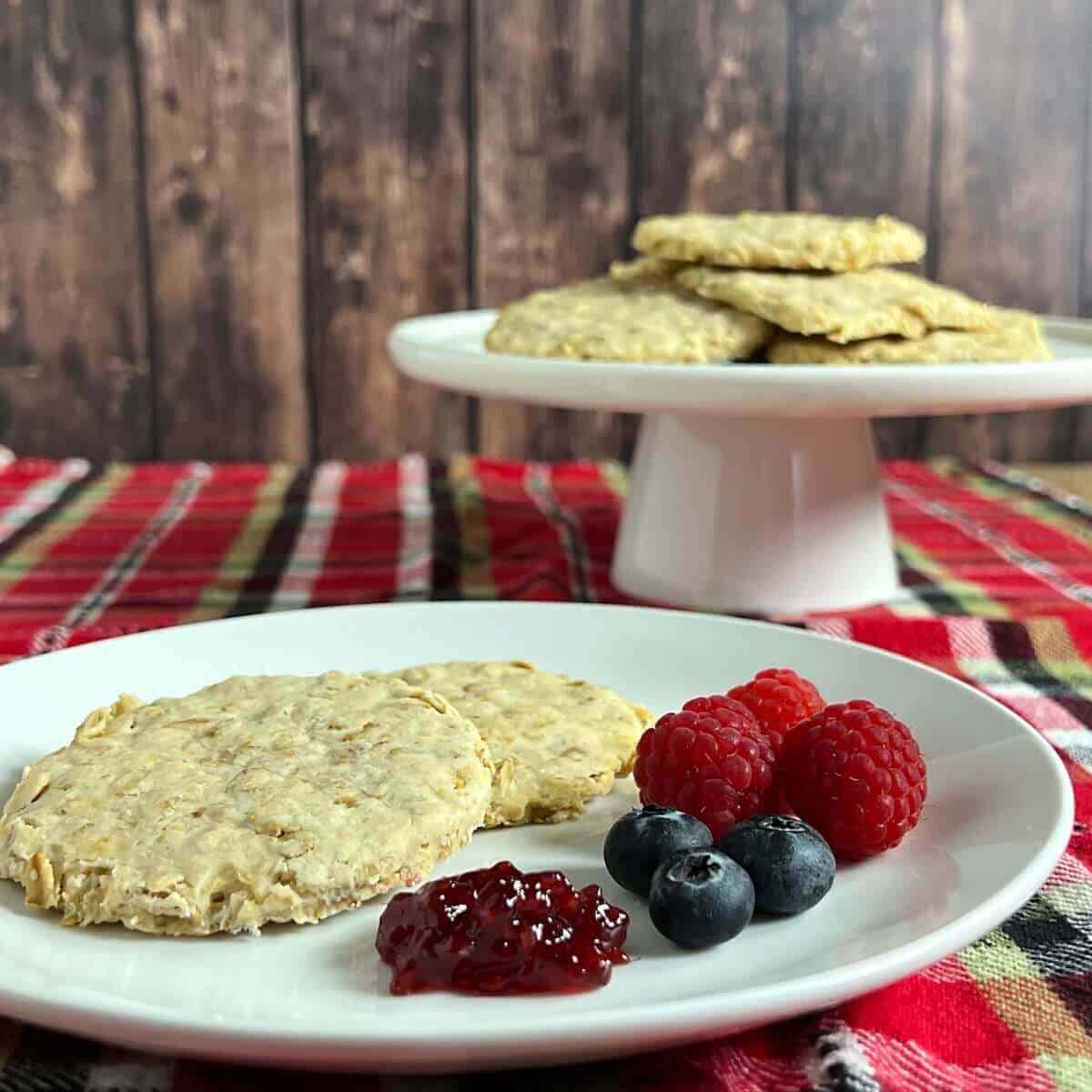 Two bannocks on a white plate with jam and berries on a red checked towel with more stacked on a cake stand behind.