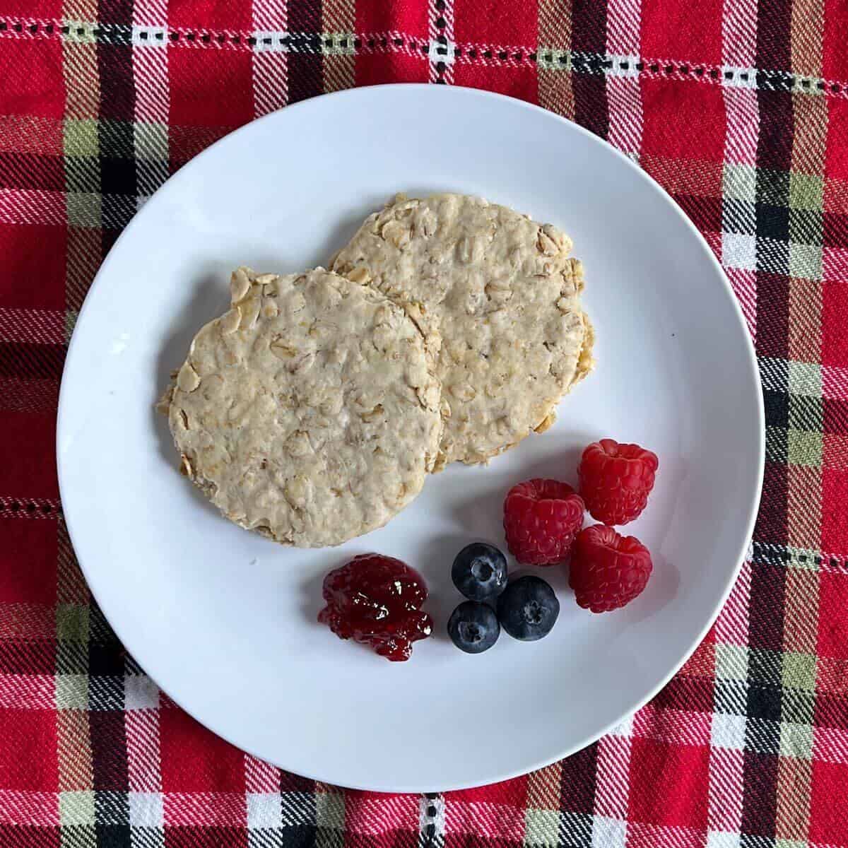 Two bannocks on a white plate with jam and berries on a red checked towel from overhead.