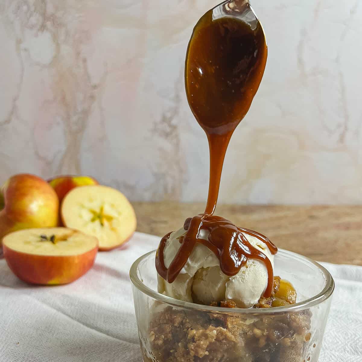 Apple Crisp in a glass bowl topped with vanilla ice cream with caramel sauce being drizzled on with apple halves behind.