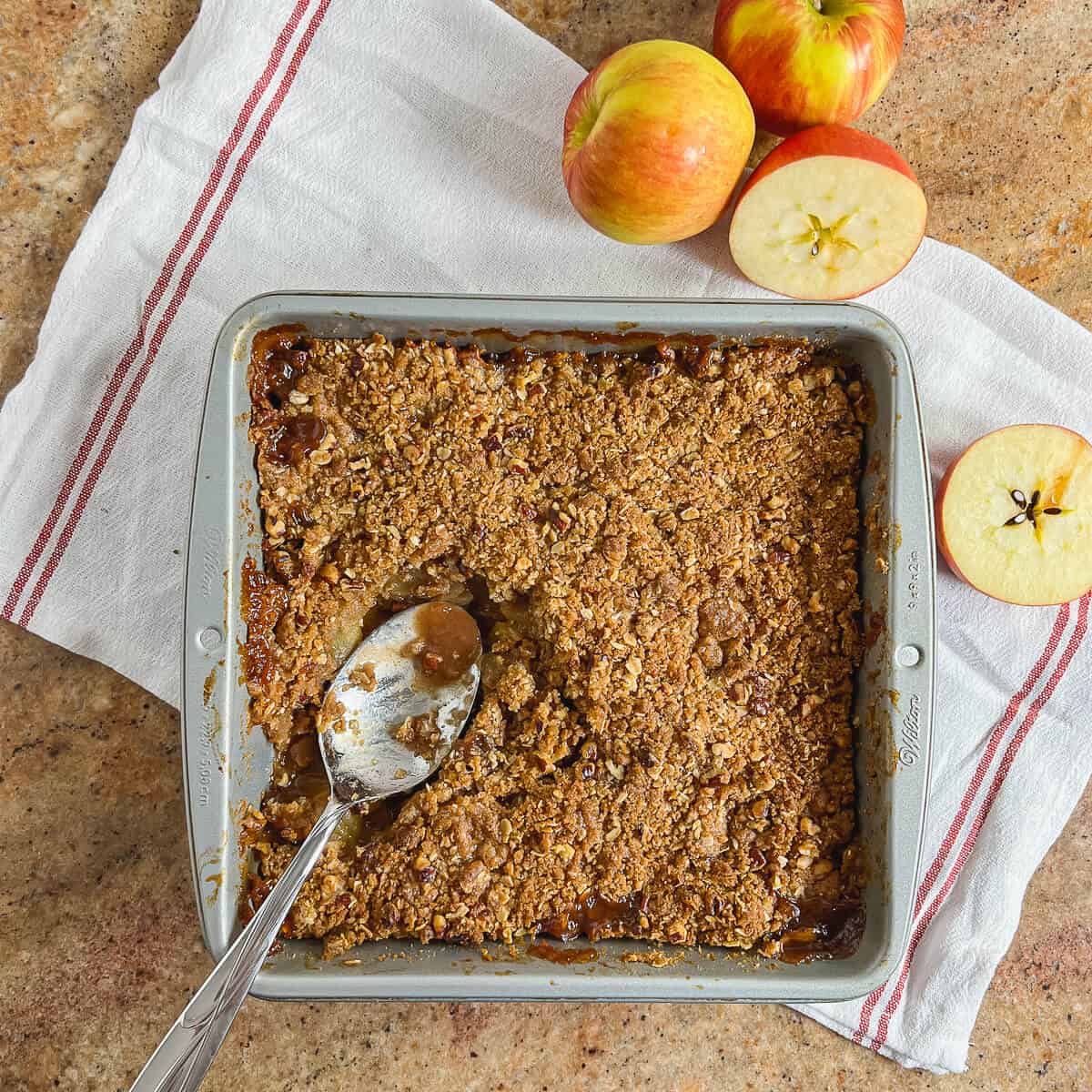 Baked Apple Crisp with a serving spoon on a white towel with red stripes with apples from overhead.