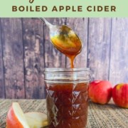 Boiled apple cider dripping from a spoon into a glass jar surrounded by apples Pinterest banner.