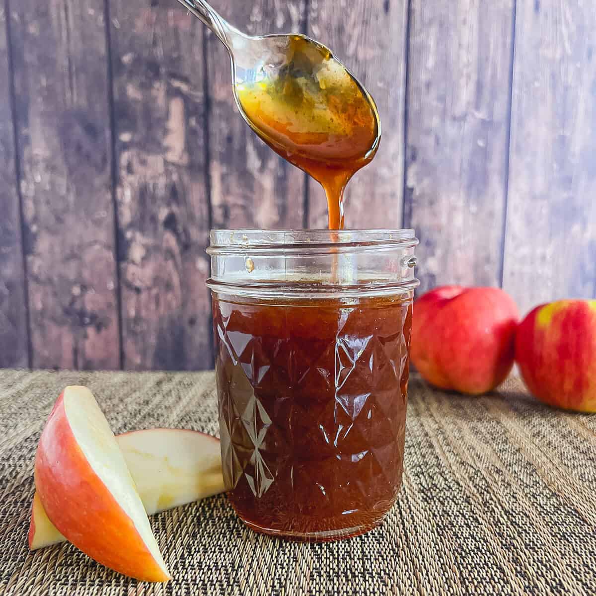 Boiled apple cider dripping from a spoon into a glass jar surrounded by apples.