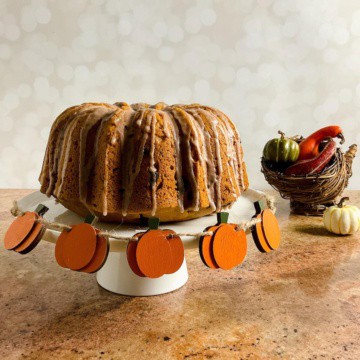 Whole sourdough pumpkin cake on a white cake stand next to a wicker cornucopia filled with gourds & small pumpkins.