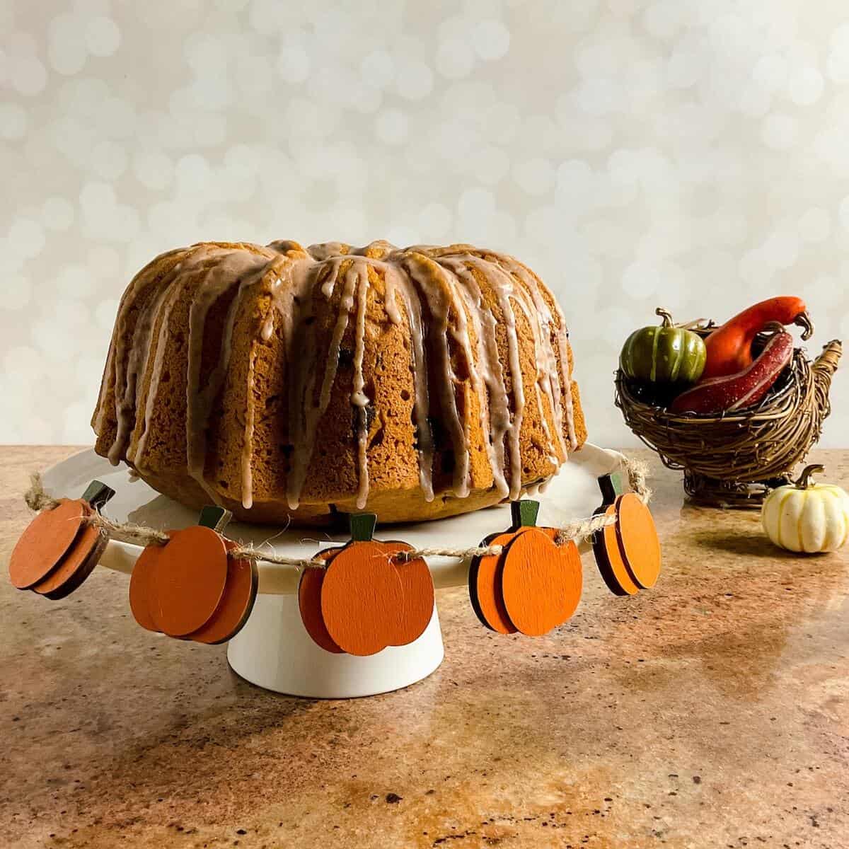 Whole sourdough pumpkin cake on a white cake stand next to a wicker cornucopia filled with gourds & small pumpkins.