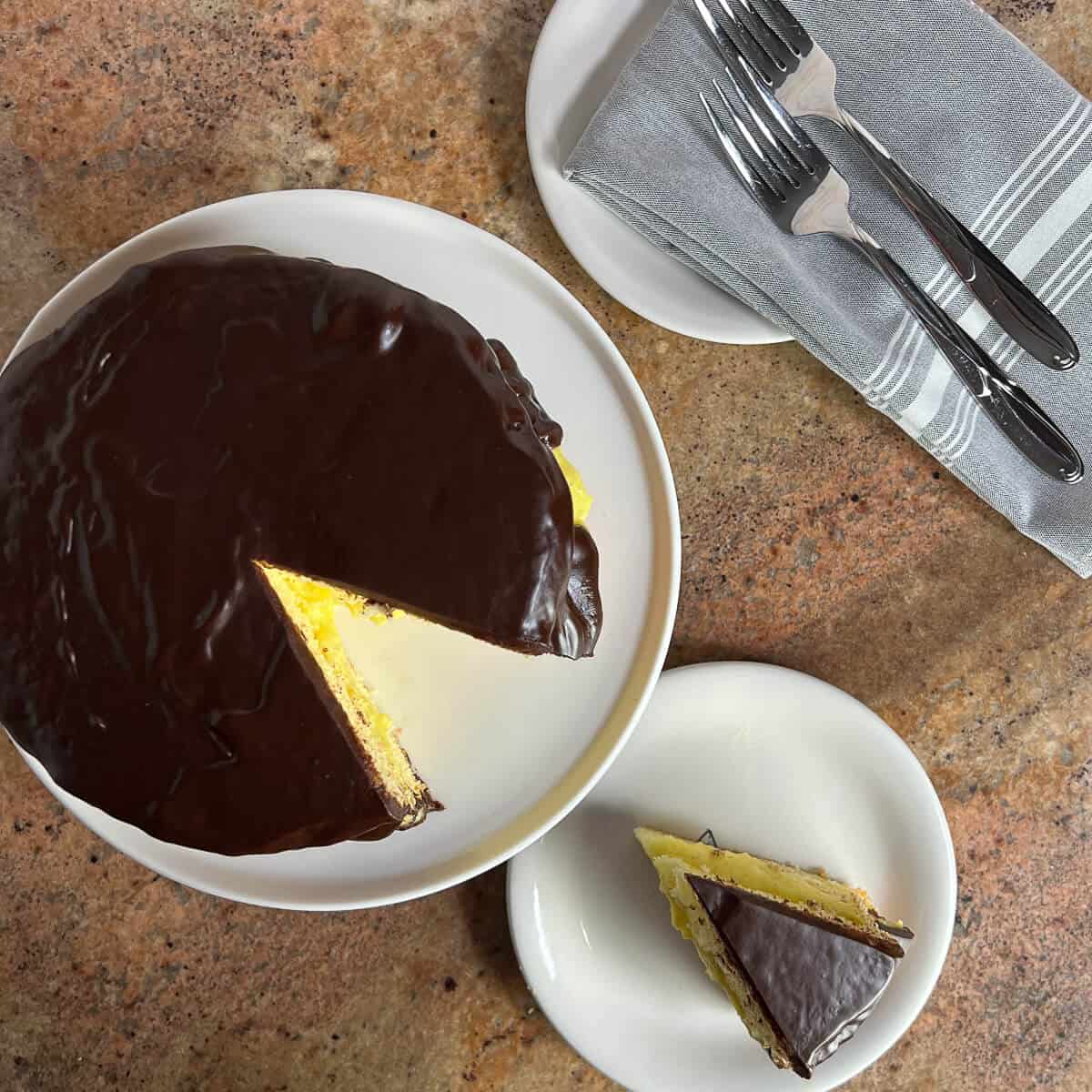 Slice of Boston Cream Pie on a white plate with the sliced whole cake on a white cake stand & 2 forks on a grey napkin from overhead..