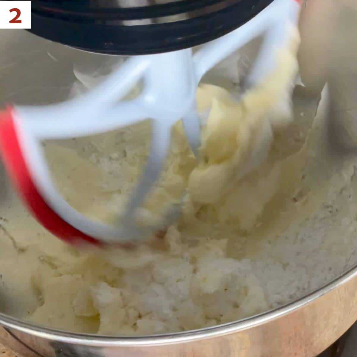 Adding powdered sugar to creamed butter in a stand mixer bowl.