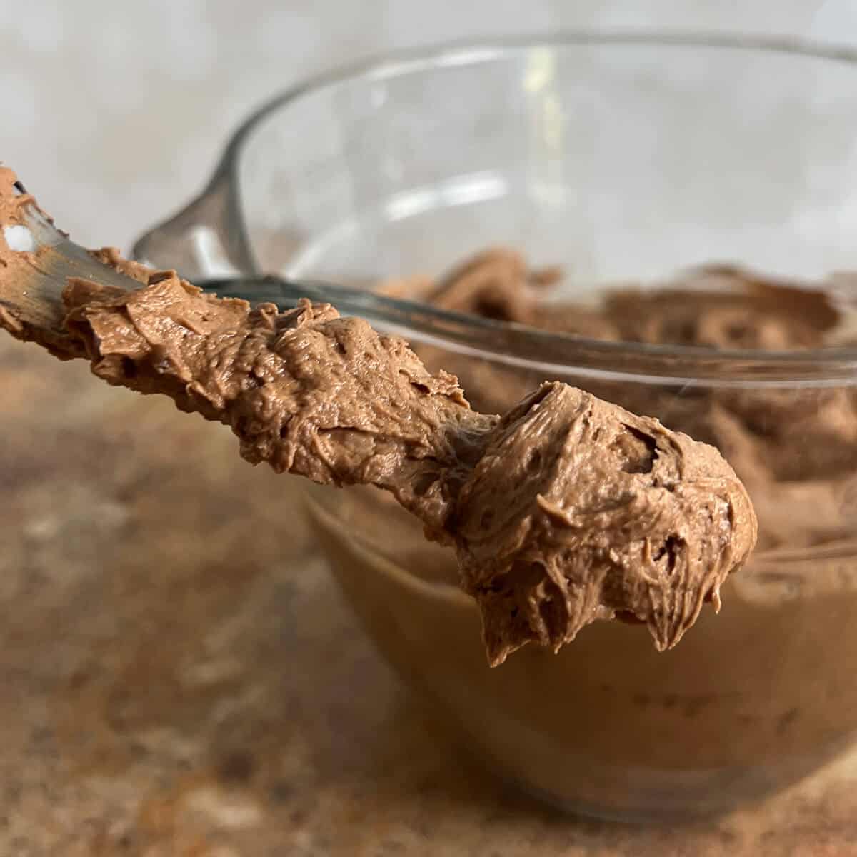 Dark chocolate buttercream frosting on a spatula with bowl of frosting behind.