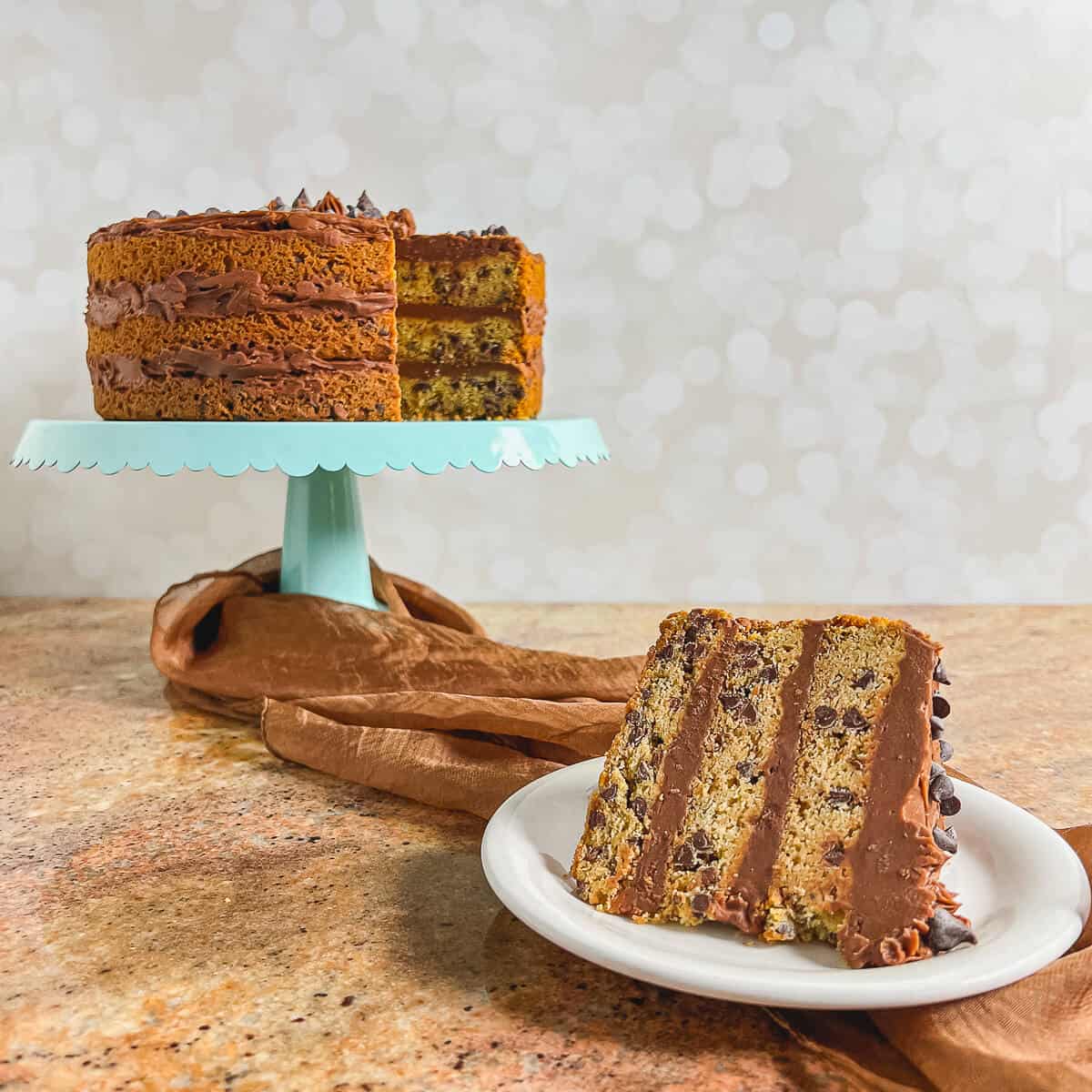 Slice of Chocolate Chip Cookie Cake on a white plate with the sliced cake on a blue cake stand behind.