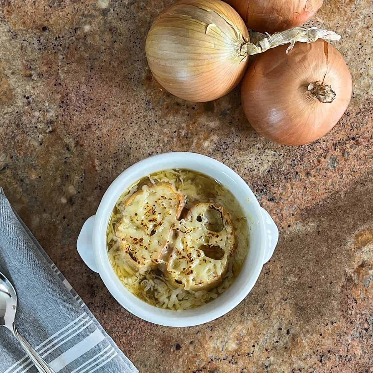 French Onion Soup in a bowl next to a spoon on a grey napkin and onions from overhead.