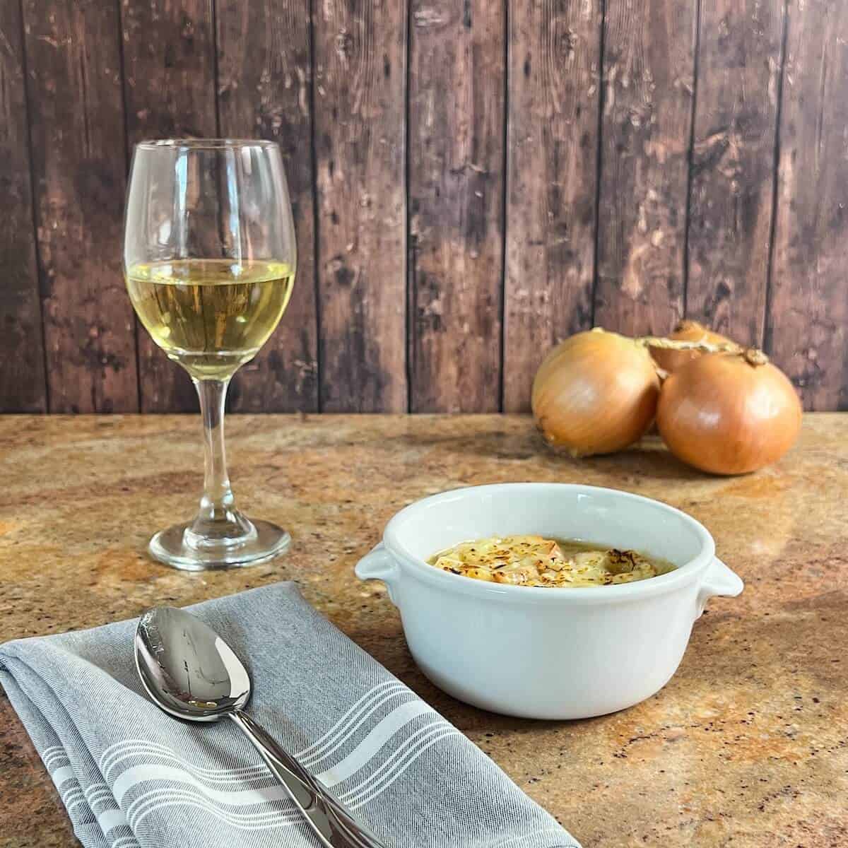 French Onion Soup in a bowl next to a spoon on a grey napkin with a glass of white wine and onions behind.