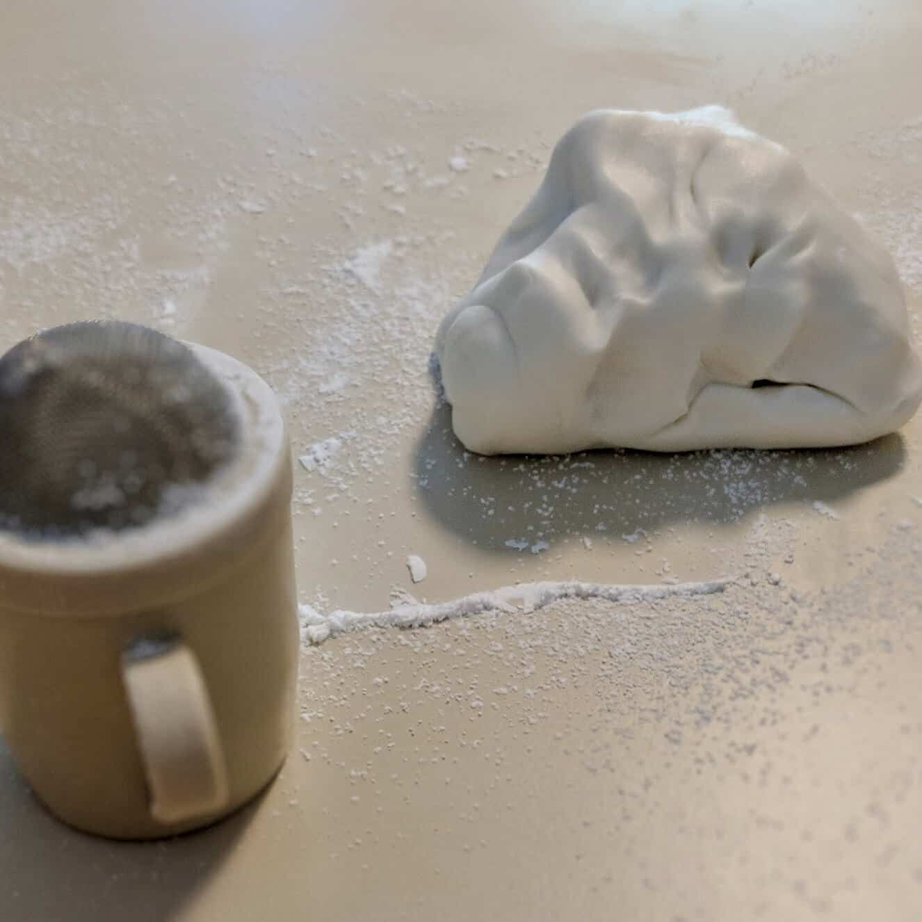 Kneaded marshmallow fondant on a powdered-sugar-dusted counter next to a shaker.