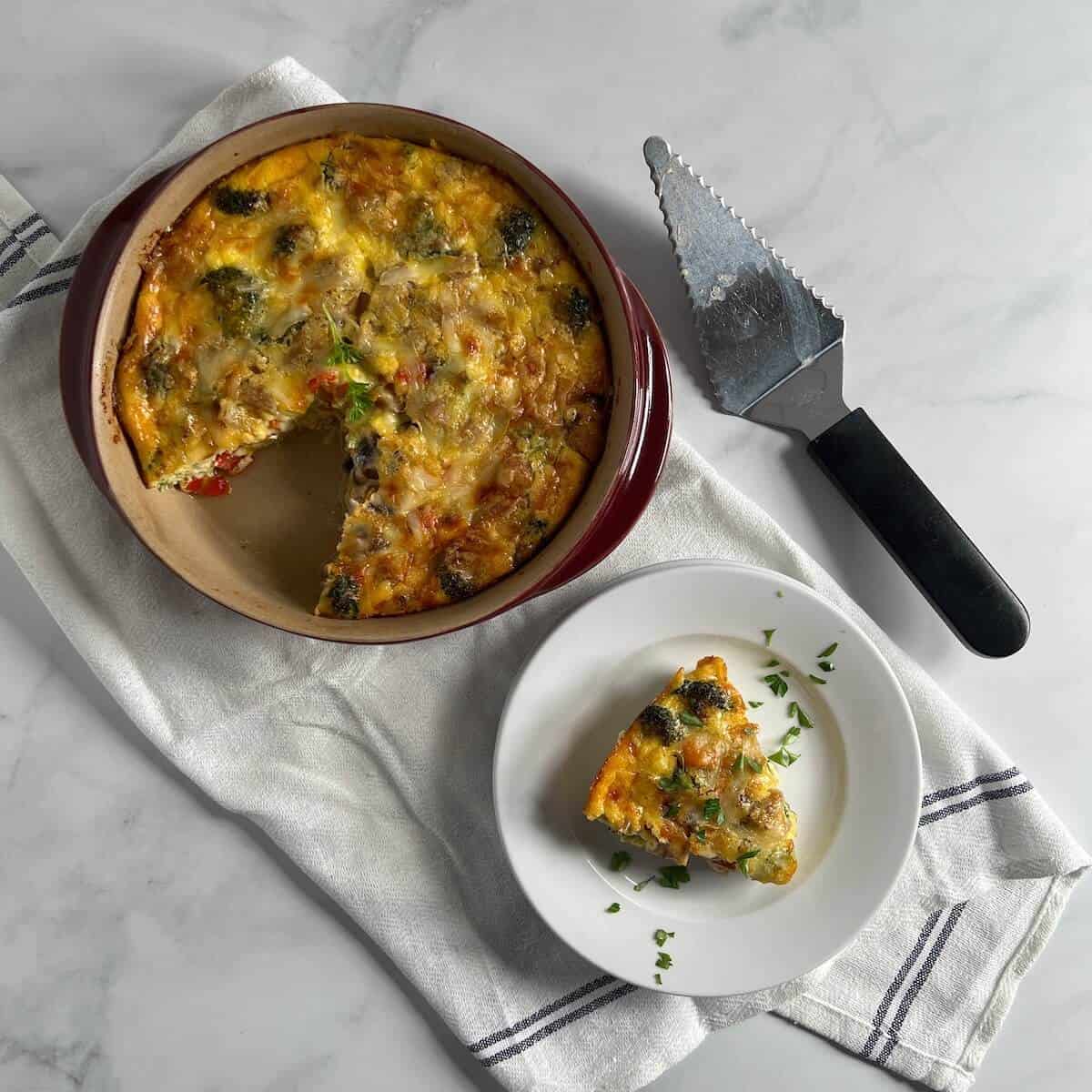 Sliced Sausage & Broccoli quiche in a red baking dish next to a slice on a round white plate and pie server on a white towel from overhead.