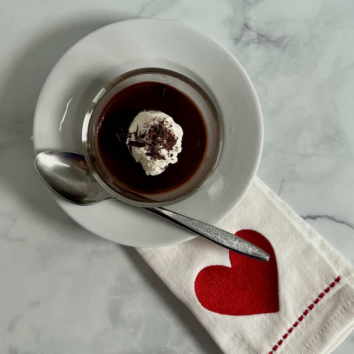 Two ramekins of pudding on round white plates with spoons next to a white towel with a red heart.