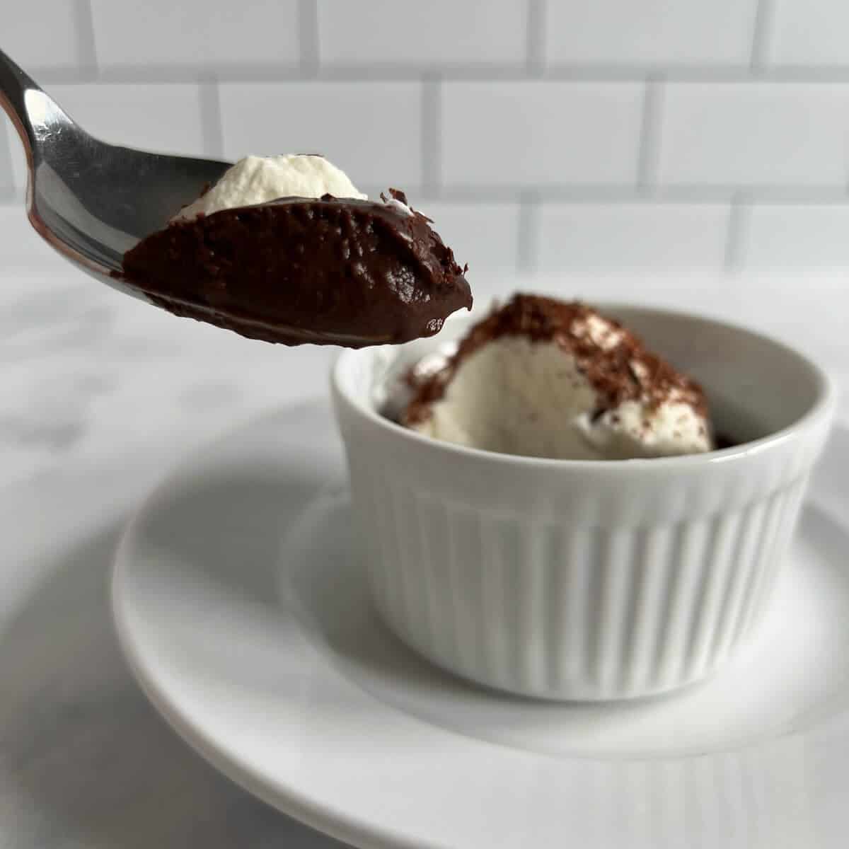 Bite of pudding on a spoon in front of a ramekin of pudding on round white plate with spoon.