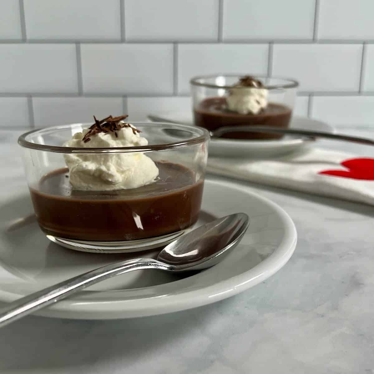 Two ramekins of pudding on round white plates with spoons next to a white towel with a red heart.