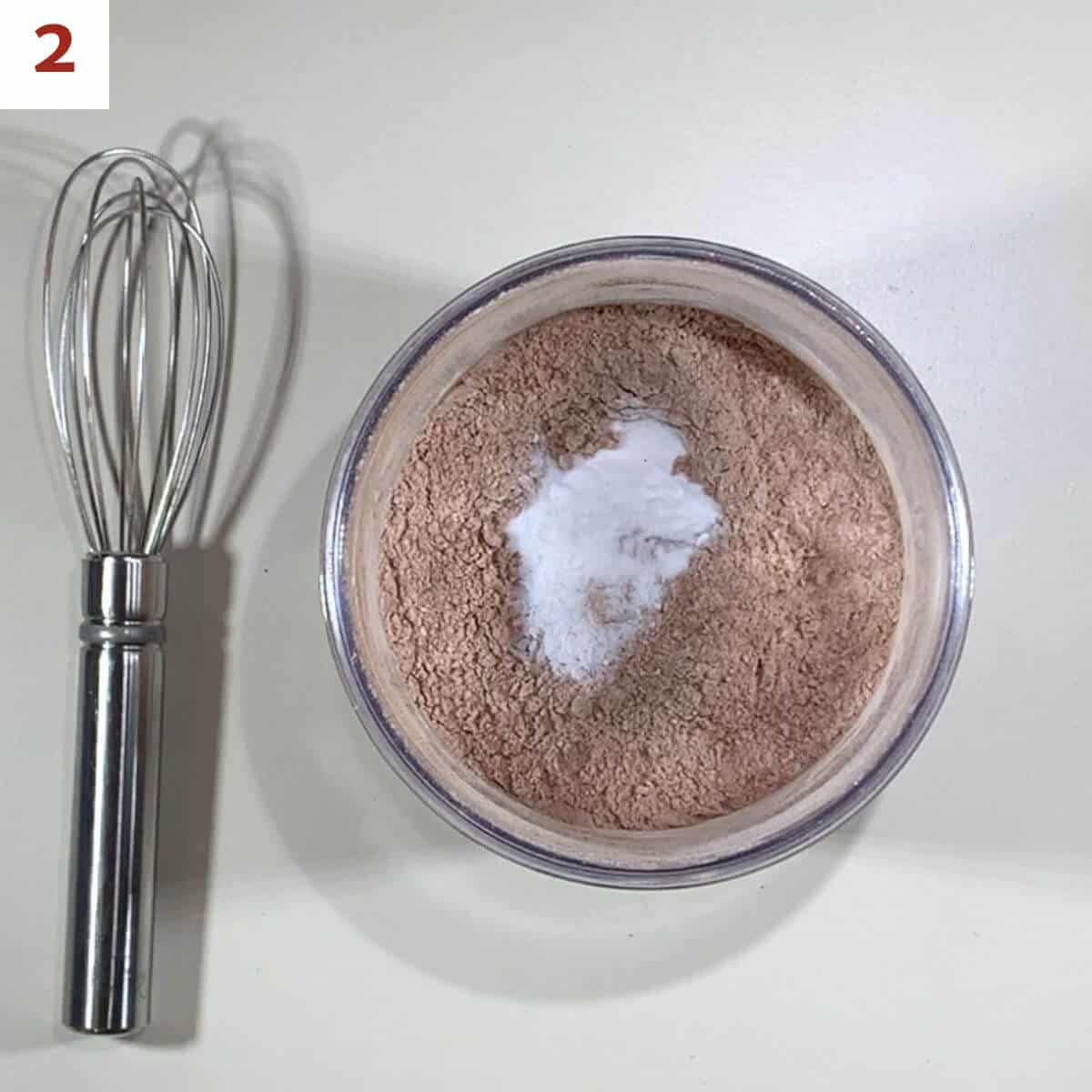 Sifted flour and cocoa powder in a small bowl topped with baking soda and salt next to a whisk.