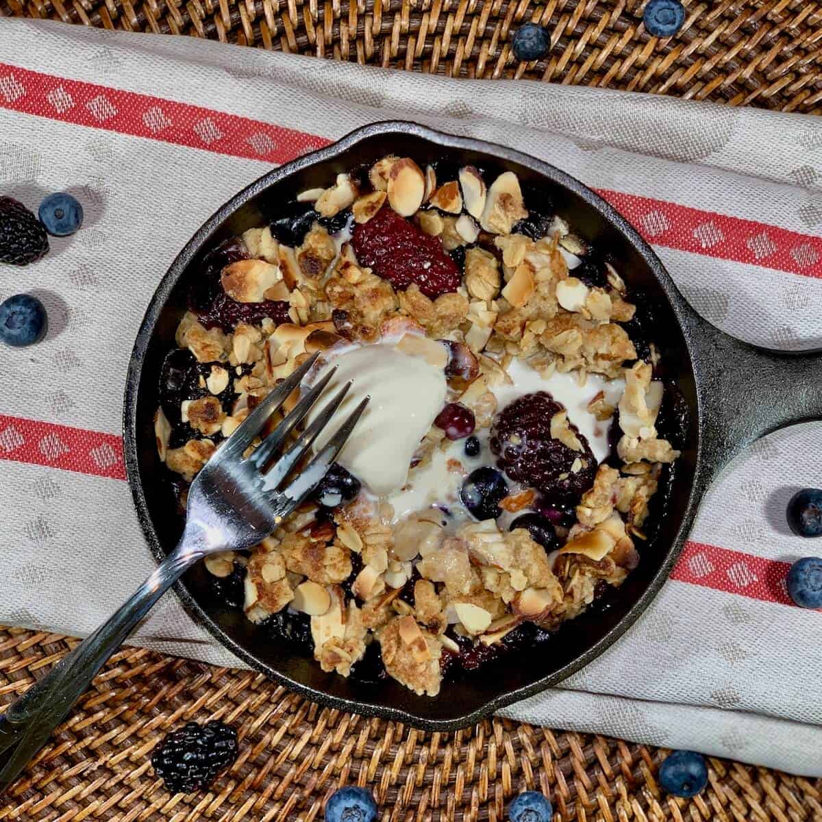 Mini Skillet Berry Crisp on a red & white striped towel with fork & scoop of vanilla ice cream surrounded by blueberries on a wooden tray from overhead.