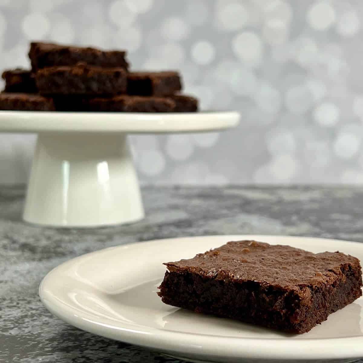 Bourbon brownies on a white plate in front of a stack of brownies on a white cake stand.