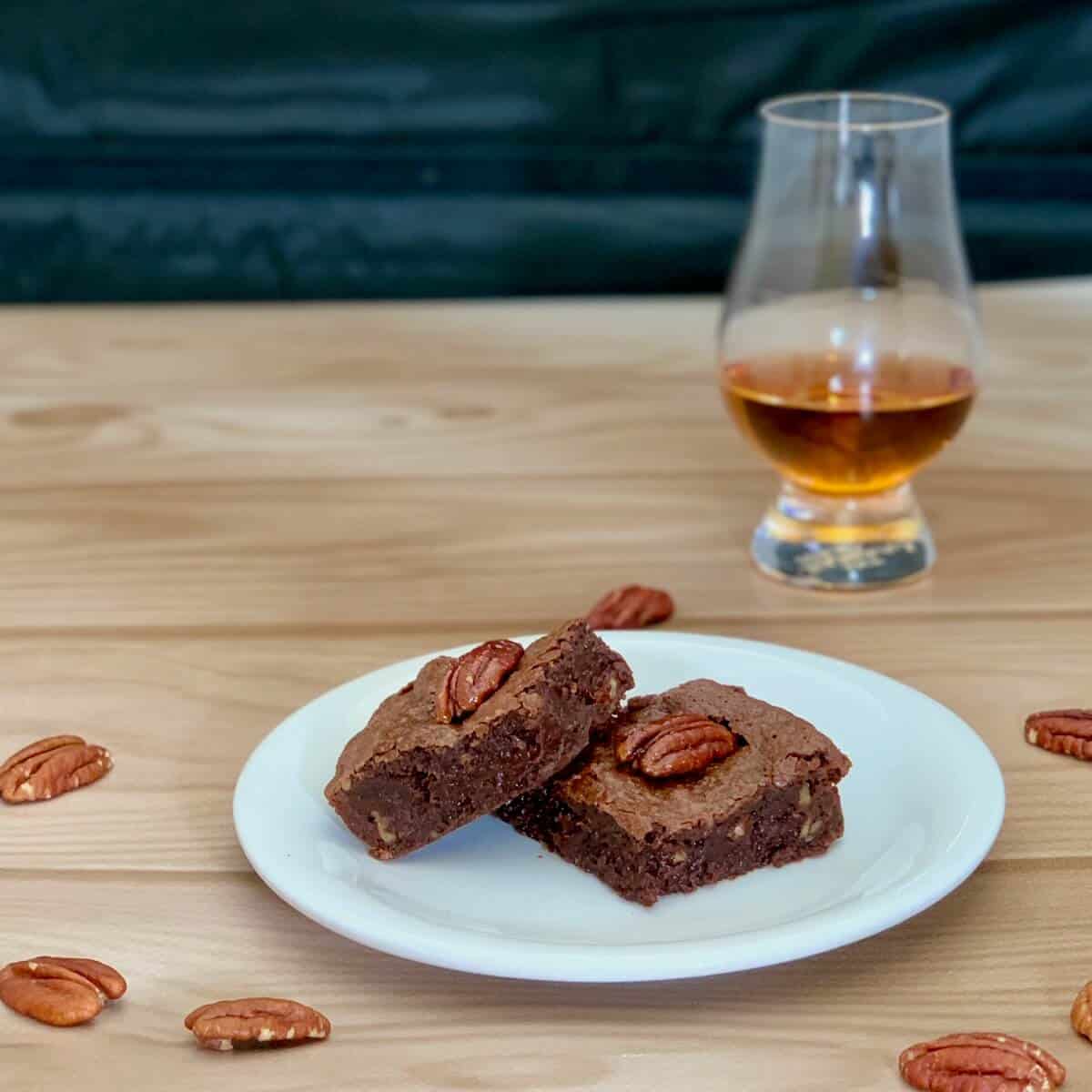 Two candied pecan whiskey browned on a white plate surrounded by whole pecans with a glass of whiskey behind.