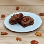 Two candied pecan bourbon brownies on a white plate surrounded by whole pecans.