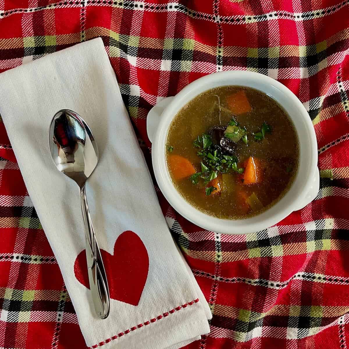 Cock-a-leekie soup in a white bowl next to a spoon on white towel with a red heart all on a red checkered towel.