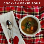 Cock-a-leekie soup in a white bowl next to a spoon on white towel with a red heart all on a red checkered towel Pinterest banner.