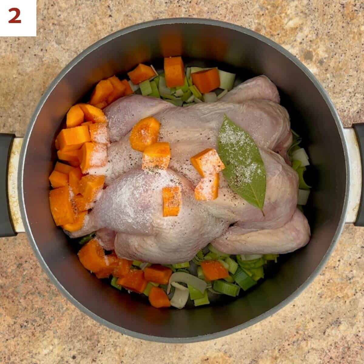 A whole chicken in a stock pot surrounded by leeks and carrots and topped with salt, pepper, and a bay leaf.