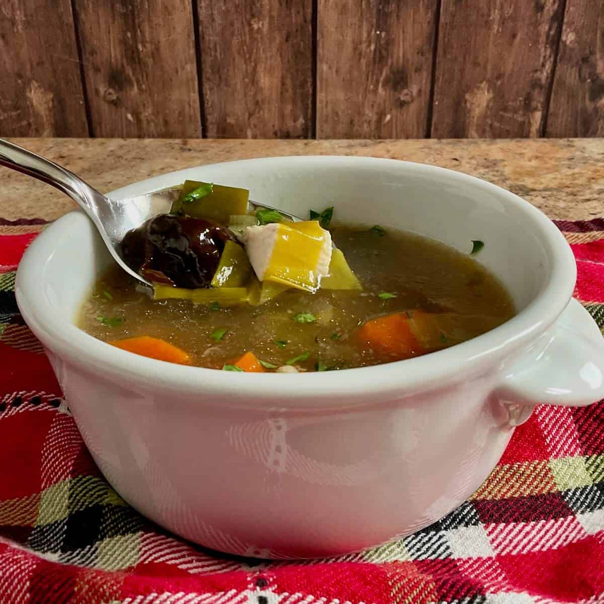 Cock-a-leekie soup in a white bowl with a spoon lifting prunes and leeks all on a red towel.