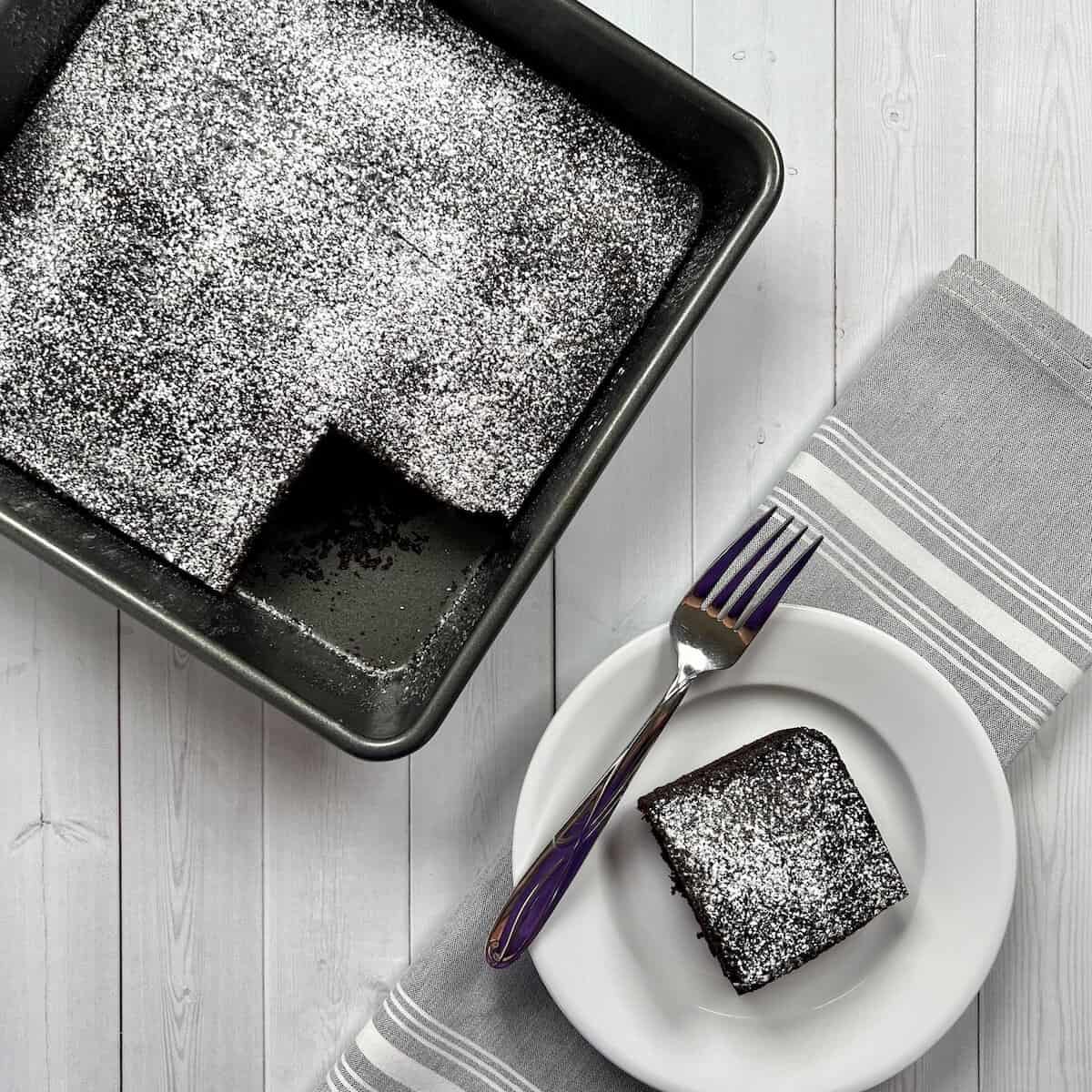 Piece of chocolate cake on a white plate next to a fork with cake in background from overhead.